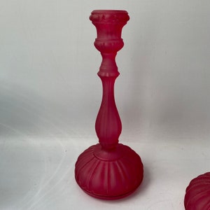 May include: A pair of frosted glass candle holders in a deep pink colour. The holders have a classic design with a fluted base and a decorative top.