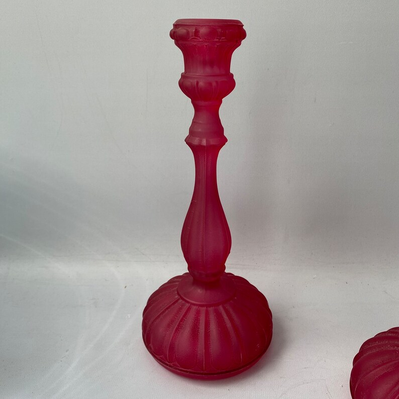 May include: A pair of frosted glass candle holders in a deep pink colour. The holders have a classic design with a fluted base and a decorative top.