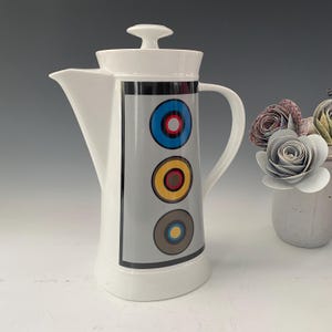 Puede incluir: Cafetera de cerámica blanca con un panel gris con tres diseños circulares coloridos. La cafetera tiene un pico, un asa y una tapa. Los círculos son azules, amarillos y marrones con detalles en rojo y negro. Un arreglo floral decorativo está en el fondo.