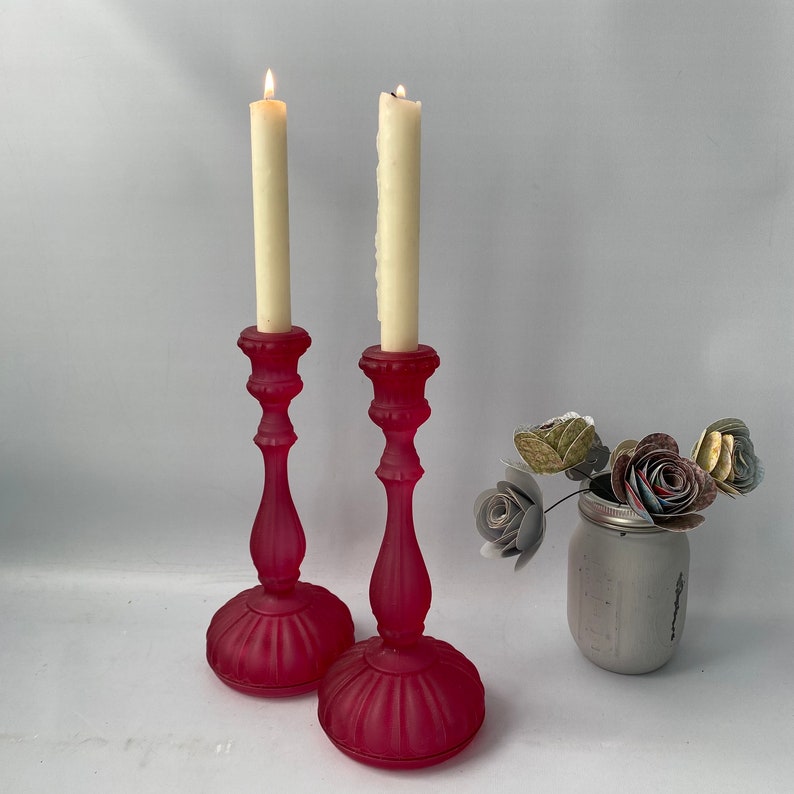 May include: Two pink glass candle holders with white candles. The candle holders have a fluted design and are on a white surface.