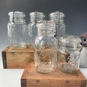 Op de afbeelding: Vijf vintage glazen potten met draadbeugels. De potten zijn van helder glas en hebben een getextureerd oppervlak. De potten zijn gelabeld met "Ball" en "Ideal".