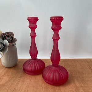May include: A pair of red glass candle holders with a frosted finish. The candle holders have a fluted base and a decorative top.