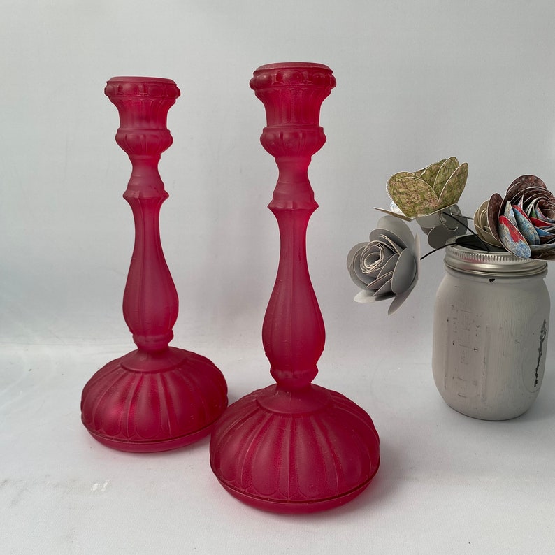May include: Two frosted glass candle holders in a deep pink colour. The candle holders have a classic design with a wide base and a tall, slender body.