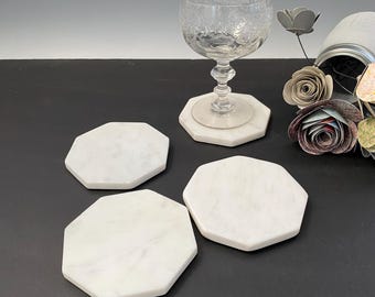 May include: Four white marble octagonal coasters on a black surface. The coasters are smooth and have a natural veining pattern.