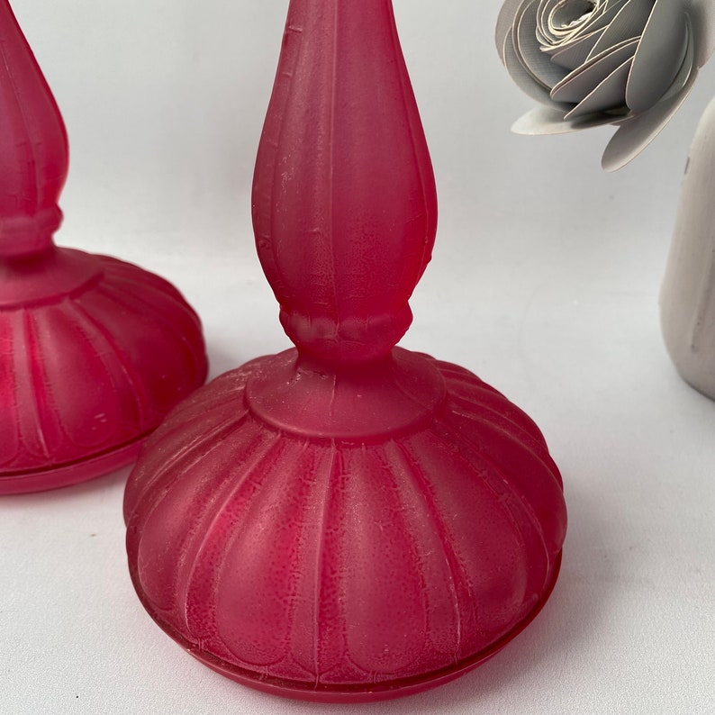 May include: Two pink frosted glass candle holders with a fluted design. The candle holders are on a white surface.