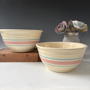 Puede incluir: Dos cuencos de cerámica color crema con rayas rosas y azules. Los cuencos están colocados sobre una superficie de madera, con un tarro de flores de papel en el fondo. Los cuencos tienen un diseño clásico y vintage.