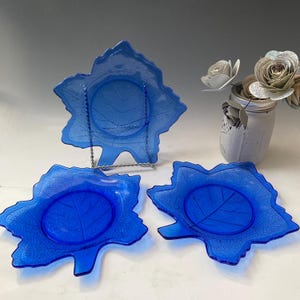 Op de afbeelding: Drie kobaltblauwe glazen borden in de vorm van esdoornbladeren. De borden hebben een reliëfdesign dat lijkt op de nerven van een blad.