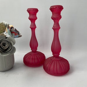 May include: Two pink glass candle holders with a frosted finish. The candle holders are tall and slender with a round base.