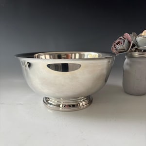 May include: A polished silver-toned metal bowl with a smooth, reflective surface and a wide, circular base. The bowl features a deep, rounded basin and a flared rim, sitting alongside a small glass jar containing decorative fabric flowers.