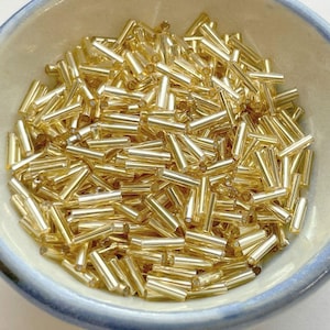 May include: A bowl filled with gold glass beads. The beads are long and thin, resembling small cylinders.