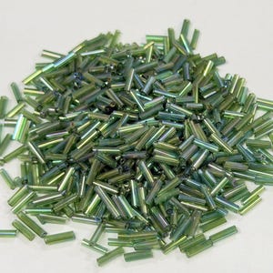 May include: A pile of iridescent green glass seed beads. These small, cylindrical beads have a metallic sheen and are scattered on a white surface. Ideal for jewelry making and crafting projects.
