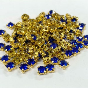 May include: A pile of blue and gold rhinestones. The rhinestones are small and have a square shape. They are perfect for adding sparkle to crafts and jewelry.