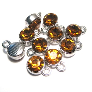 May include: A group of ten silver-toned metal charms with amber-colored glass rhinestones. Each charm has a loop for attaching to a necklace, bracelet, or other jewelry.