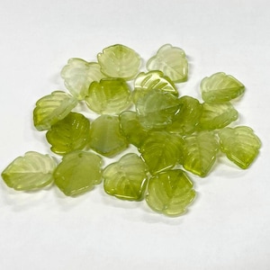 May include: A collection of translucent green leaf-shaped glass beads. The beads vary in shades of green, from light to medium, and have detailed veining. These decorative beads are ideal for jewelry making or craft projects.