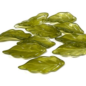 May include: A collection of olive-green glass leaf-shaped beads. Each bead has a hole for stringing and a textured surface, resembling real leaves. The beads are arranged on a white background.