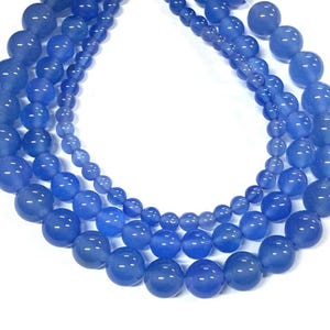 May include: Three strands of blue gemstone beads in varying sizes. The beads are a solid, light blue color with a glossy finish. The strands are arranged in a layered pattern, showcasing the different bead sizes and the overall design.