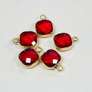 May include: Five red square-cut gemstone charms with gold-colored metal settings. Each charm has a loop for attaching to jewelry. The gems are a deep, vibrant red color, and the gold settings add a touch of elegance.