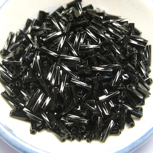 May include: A bowl filled with black glass beads. The beads are long and narrow, with a faceted finish.