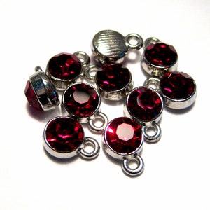 May include: A group of ten red rhinestone charms with silver settings and loops. The rhinestones are round and have a faceted design.
