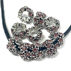 May include: A necklace with a black cord and silver-toned floral accents. The floral accents are adorned with small, pink gemstones. Several loose floral accents are also present, creating a set.