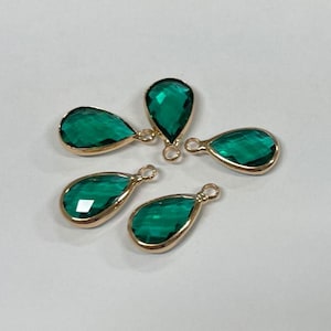 May include: Five teardrop-shaped green gemstone charms with gold-colored metal edging. Each charm has a small loop at the top for attaching to jewelry. The gemstones are faceted, reflecting light.