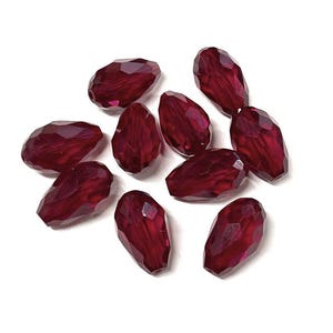 May include: A collection of faceted, teardrop-shaped beads in a deep burgundy color. The beads have a glossy, reflective surface and are scattered against a white background. These beads are suitable for jewelry making.
