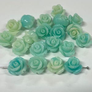 May include: A collection of 18 light blue and green plastic rose flower beads. The beads are in various shades of blue and green, with some having a white center.