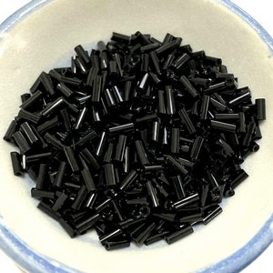 May include: A bowl filled with black glass seed beads. The beads are cylindrical in shape and have a glossy finish.
