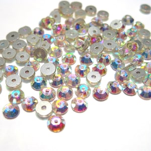 May include: A large group of iridescent clear glass rhinestones with silver settings. The rhinestones are scattered on a white surface.