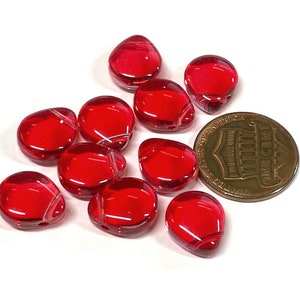 May include: A group of ten red glass beads, each with a teardrop shape and a slightly translucent appearance. The beads are scattered on a white surface, with a US penny for scale.