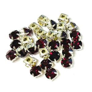 May include: A collection of sparkling, faceted, dark red rhinestones with silver-colored metal settings. The rhinestones are round and square-shaped, scattered against a white background. These embellishments are ideal for adding sparkle to crafts and accessories.