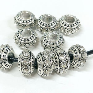 May include: Silver tone round beads with clear rhinestones. The beads have a decorative design around the edge and a hole in the center. The beads are strung on a black cord.