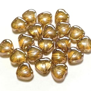 May include: A group of 20 gold heart-shaped glass beads with a shimmery finish. The beads are scattered on a white surface.