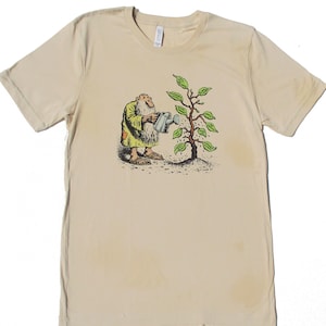以下が含まれることがあります： ベージュのTシャツに、植物に水をやる男性の漫画のイラストが描かれています。男性は緑色のローブを着て、ジョウロを持っています。植物は緑の葉と茶色の茎を持っています。