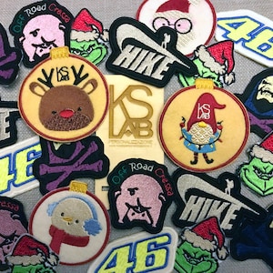 Op de afbeelding: Een verzameling kleurrijke geborduurde patches met verschillende ontwerpen, waaronder een rendier, een gnome, een Grinch, een schedel en gekruiste beenderen, en het woord "HIKE".