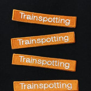 Può includere: Quattro toppe di tessuto arancione con testo ricamato bianco che recita "Trainspotting".