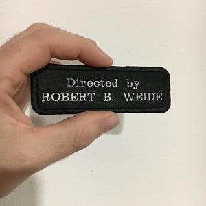 Può includere: Toppa in tessuto nero con ricamo bianco che recita "Directed by ROBERT B. WEIDE"