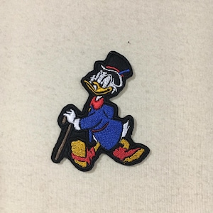 Könnte beinhalten: Ein gestickter Cartoon-Patch von Dagobert Duck, einer Disney-Figur, der einen Zylinder trägt und einen Stock hält. Er geht mit dem rechten Fuß nach vorne und dem linken Fuß nach hinten. Der Patch ist schwarz, blau, gelb, rot und weiß.