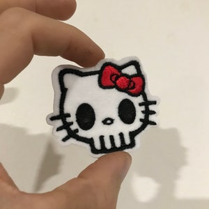 Peut inclure: Patch thermocollant brodé blanc représentant un crâne avec un nœud rouge, ressemblant à un personnage Hello Kitty.