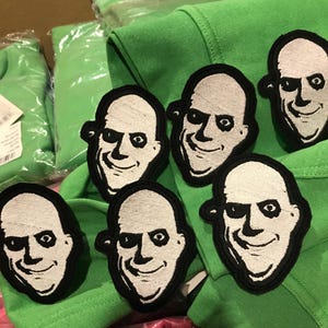 Könnte beinhalten: Sechs schwarze und weiße gestickte Bügeleisen-Aufnäher eines kahlköpfigen Mannes mit einem unheimlichen Lächeln. Die Aufnäher befinden sich auf einem grünen Hintergrund.