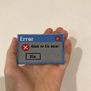 Op de afbeelding: Geborduurde patch met een computerfoutmelding. De patch heeft een blauwe achtergrond met het woord "Error" bovenaan en de tekst "Click to fix error" eronder. Er is een rode "X" aanwezig. De patch heeft ook een "Fix"-knop.