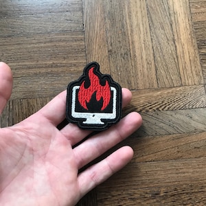 Puede incluir: Parche bordado en blanco y negro con un monitor de ordenador blanco y llamas rojas. El parche se sostiene en una mano. El fondo es una superficie de madera.