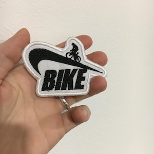 Puede incluir: Parche bordado con la palabra "BIKE" en negro, con un gráfico de ciclista. El parche tiene un borde blanco y un diseño de swoosh negro. El parche se sostiene en una mano.