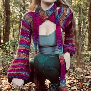 Puede incluir: Un top de crochet a rayas de colores con mangas largas y escote cruzado. El top se lleva sobre un top de tirantes gris y leggings negros.