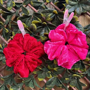 May include: Two velvet scrunchies, one red and one pink, with plastic clips attached. The scrunchies are displayed against a green leafy background.