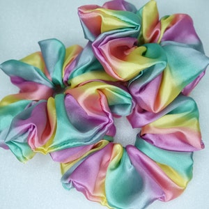May include: A set of three colorful hair scrunchies made of satin fabric. The scrunchies are in shades of pink, yellow, green, and blue, creating a rainbow effect.