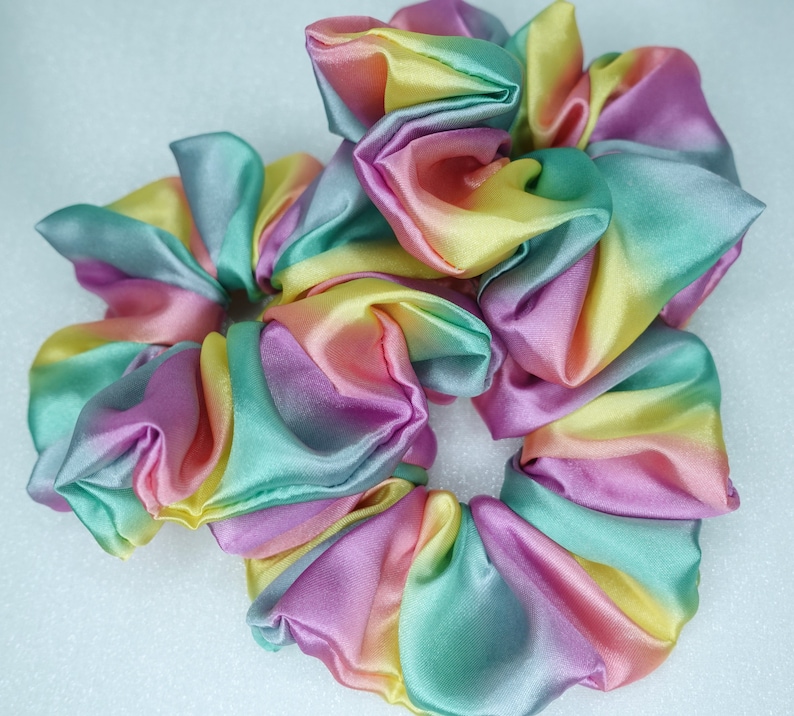 May include: A set of three colorful hair scrunchies made of satin fabric. The scrunchies are in shades of pink, yellow, green, and blue, creating a rainbow effect.