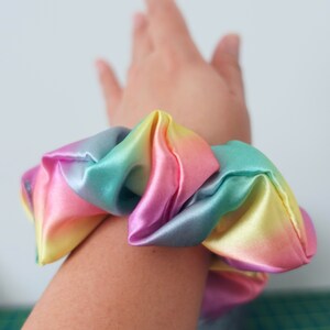 May include: A rainbow-colored scrunchie made of satin fabric. The scrunchie is tied around a person's wrist.