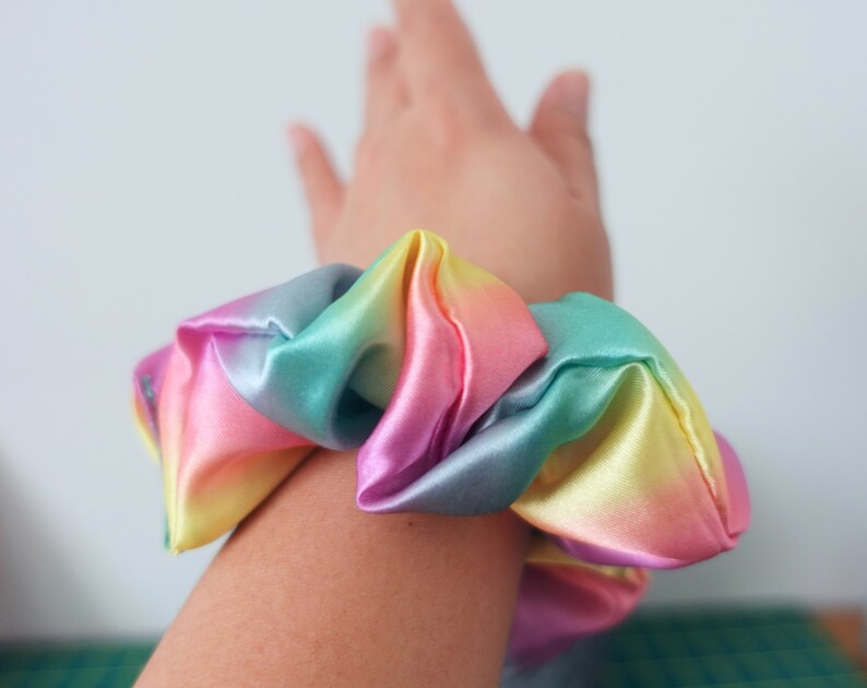 May include: A rainbow-colored scrunchie made of satin fabric. The scrunchie is tied around a person's wrist.
