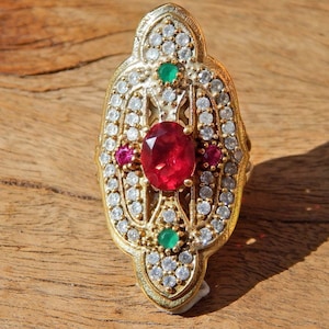 Peut inclure: Une bague en or avec une grande pierre rouge entourée de petites pierres blanches et roses. La bague a un design détaillé avec une pierre verte au sommet.